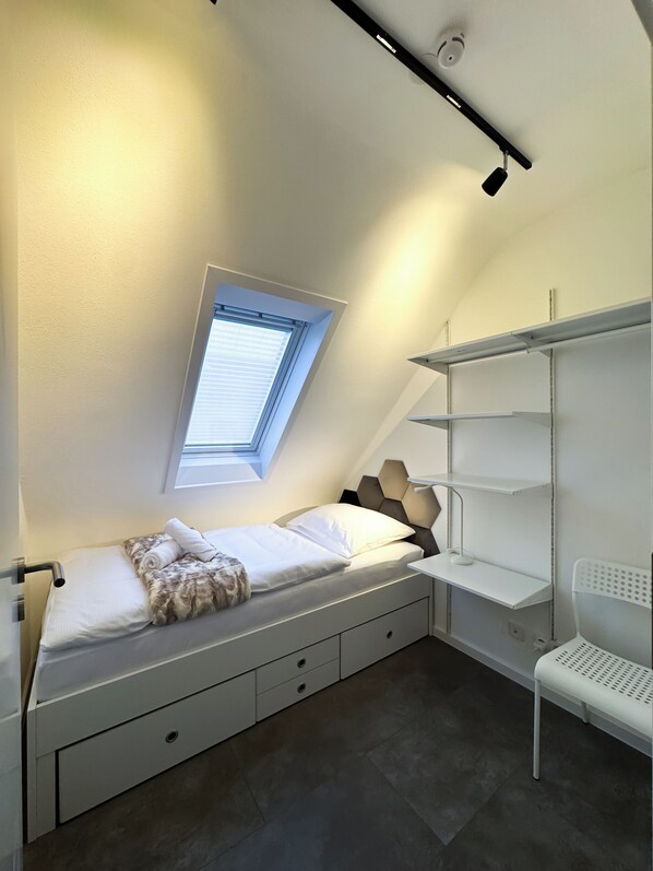Un dormitorio pequeño y moderno con tragaluz, cama con cabecera, escritorio blanco y silla blanca.