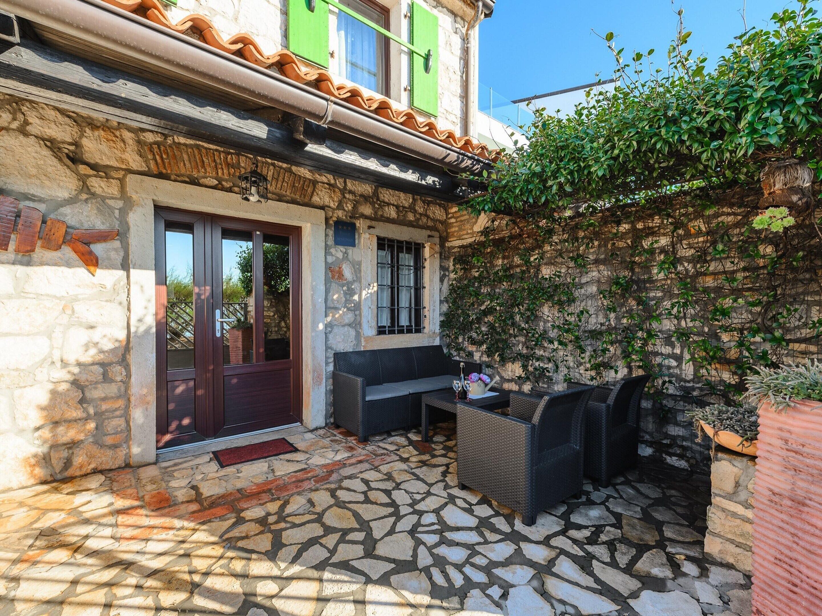 A stone-floored patio with wicker furniture and a small table, a door, and a wall with climbing plants.
