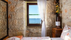 Un dormitorio con pared de piedra, ventana con vista al mar, cama de madera, mesita de noche con lámpara y un jarrón con girasoles.