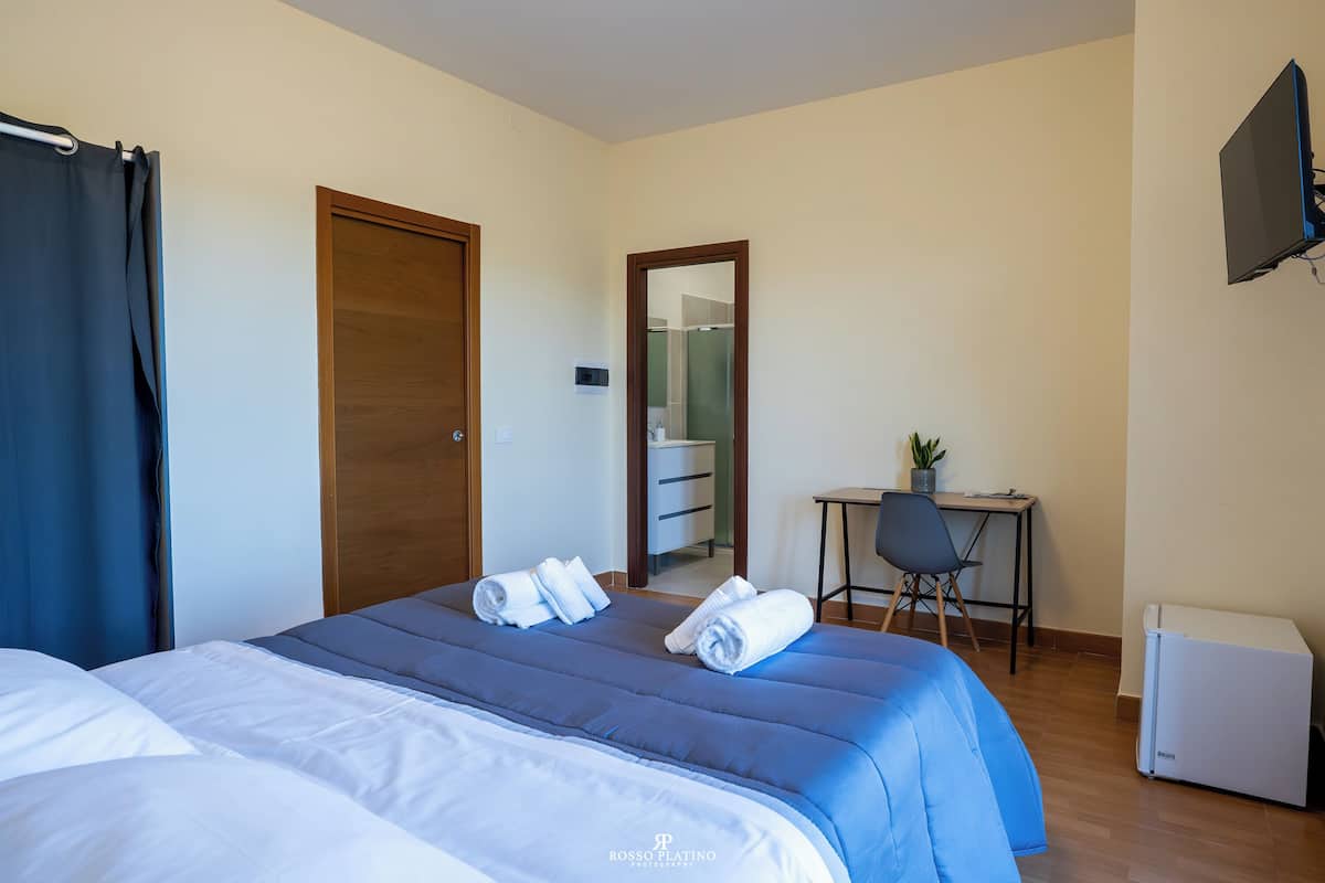 A hotel room with two beds, a desk, a chair, a small table with a plant, and a TV mounted on the wall.