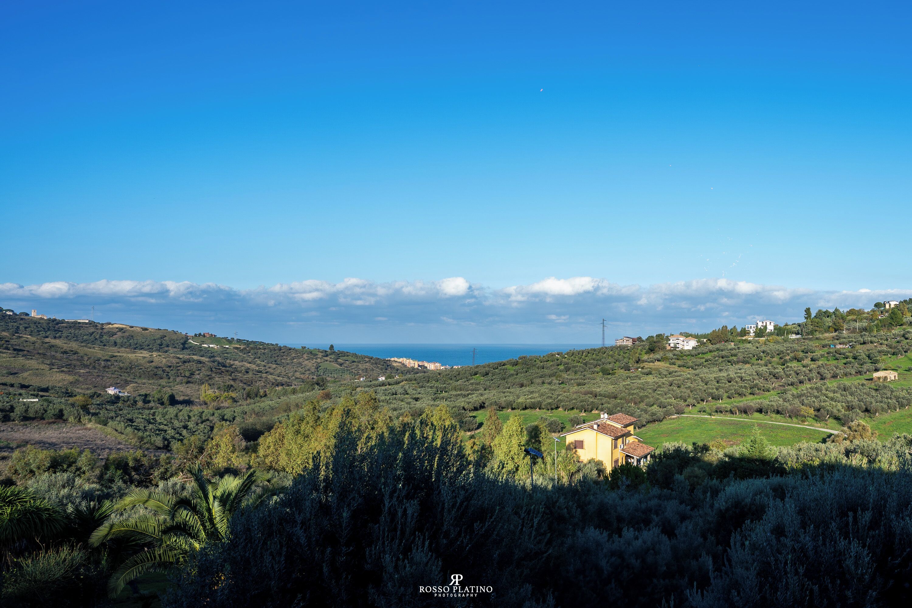 Paisaje rural con colinas ondulantes, una casa pequeña con techo rojo y cielo azul despejado.