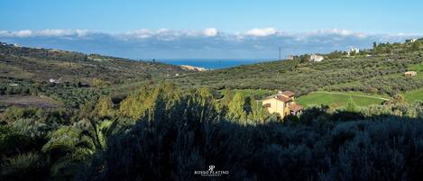 Paisaje rural con colinas ondulantes, una casa pequeña con techo rojo y cielo azul despejado.