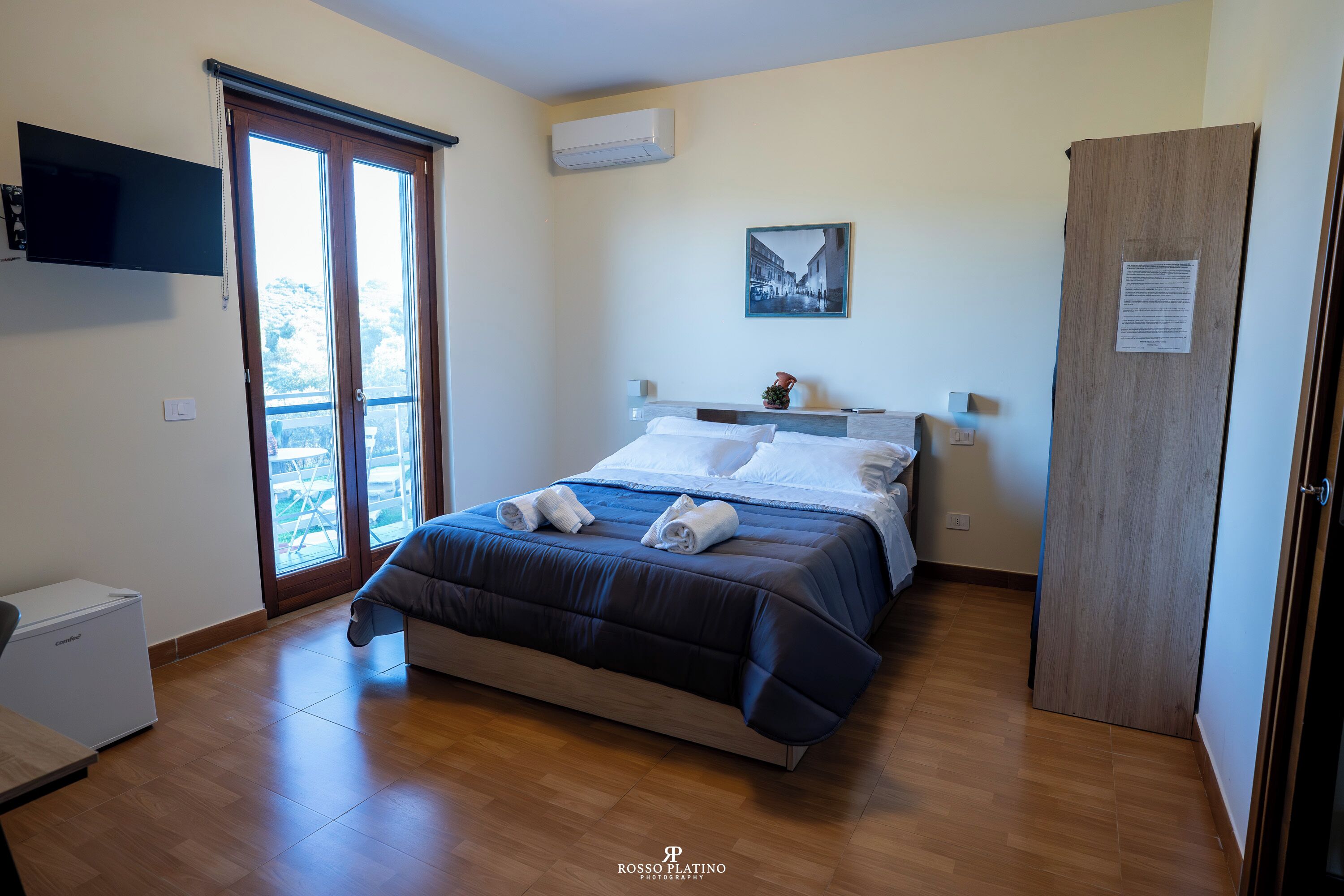 Habitación de hotel con cama, televisor de pantalla plana, armario de madera y puerta corredera de vidrio al balcón.