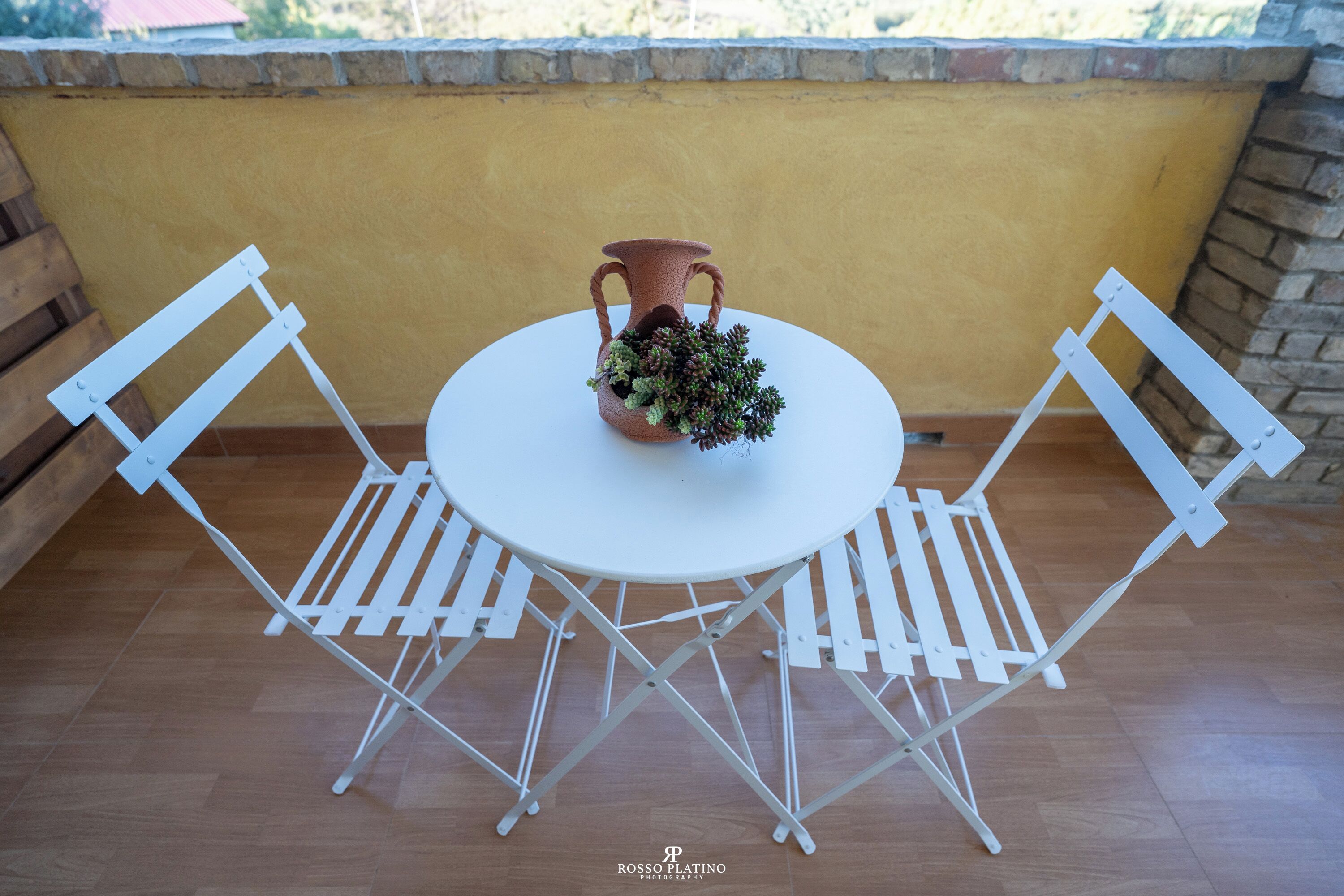 Une table ronde avec deux chaises blanches, un vase en terre cuite orné de verdure et, en arrière-plan, un mur jaune.