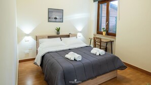 Ein ordentlich bezogenes Bett mit dunkler Bettdecke, zwei Nachttischlampen, ein kleiner Tisch mit Stuhl und ein Fenster mit Blick ins Freie.