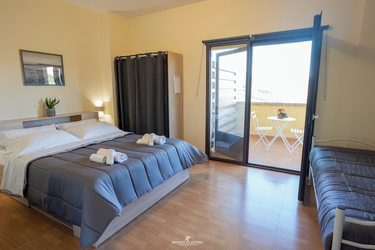 A hotel room with a bed, a small table with chairs, a sliding glass door to an outdoor area, and a potted plant.