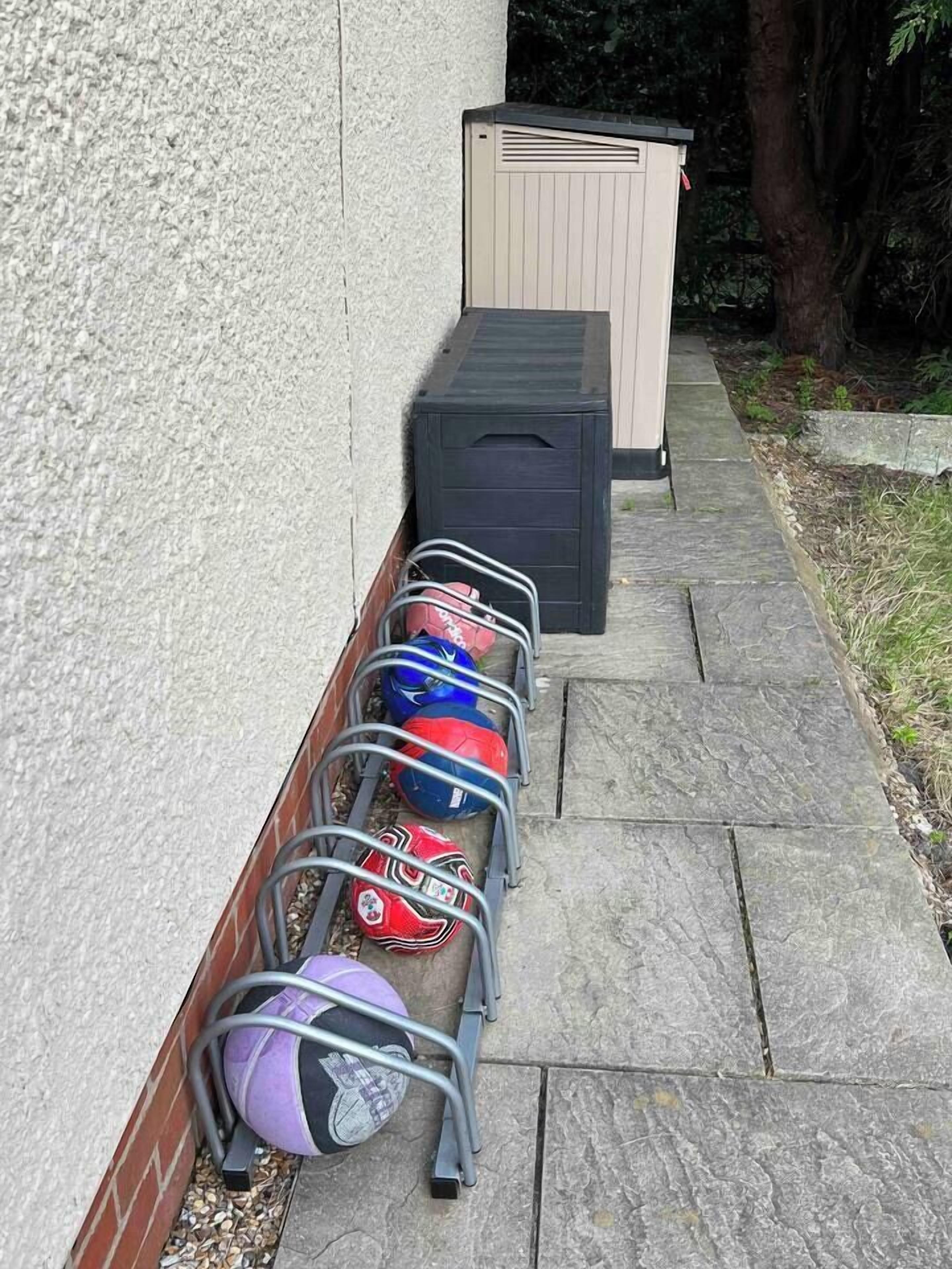 A metal rack with sports balls against a wall, a black storage box, and a concrete pathway.