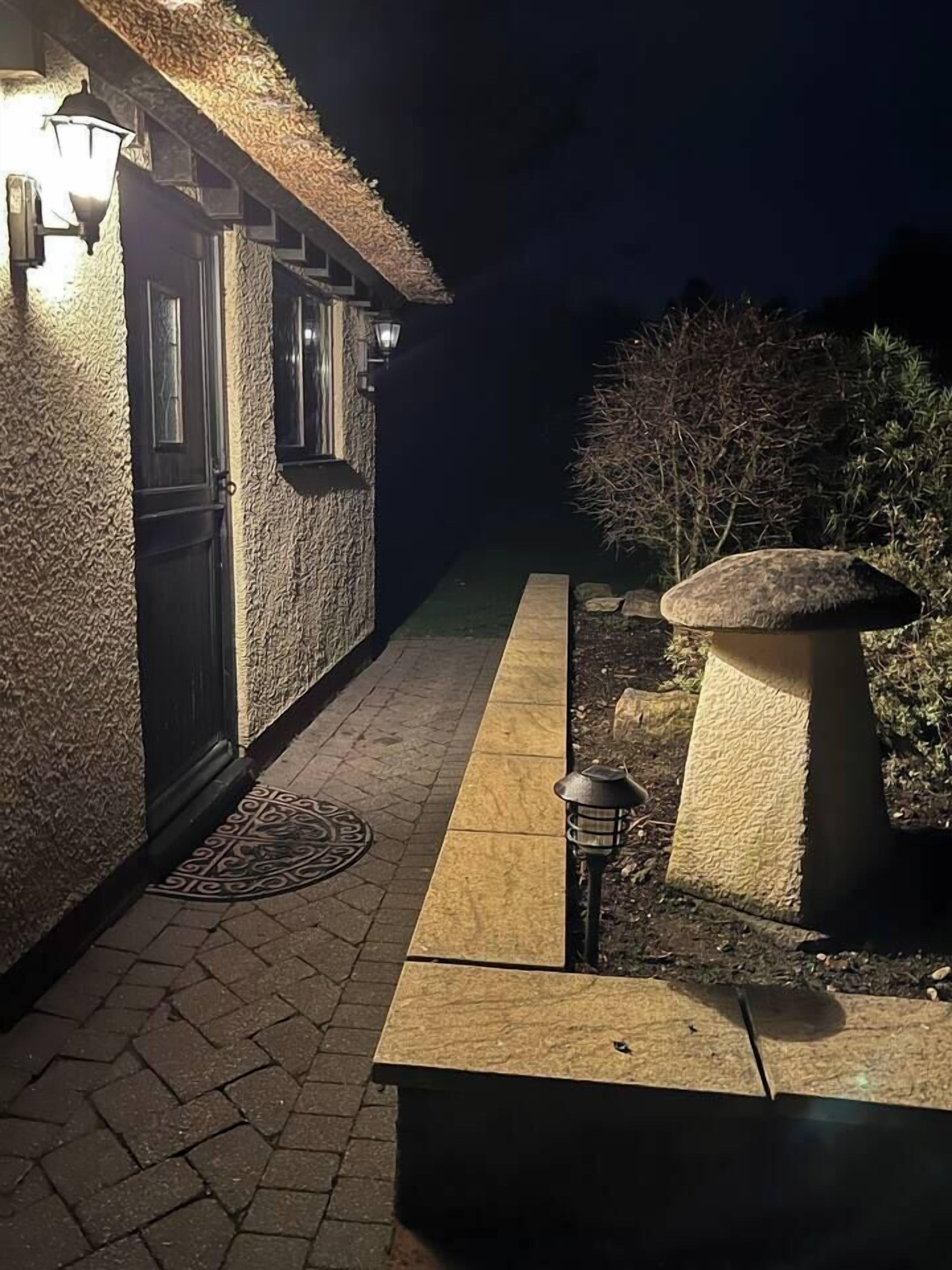 Un chemin mène à un bâtiment dont l’extérieur présente une texture particulière et qui est surmonté d’un toit de chaume. Deux lampes sont fixées au mur et un pot de fleurs se trouve près de l’entrée.