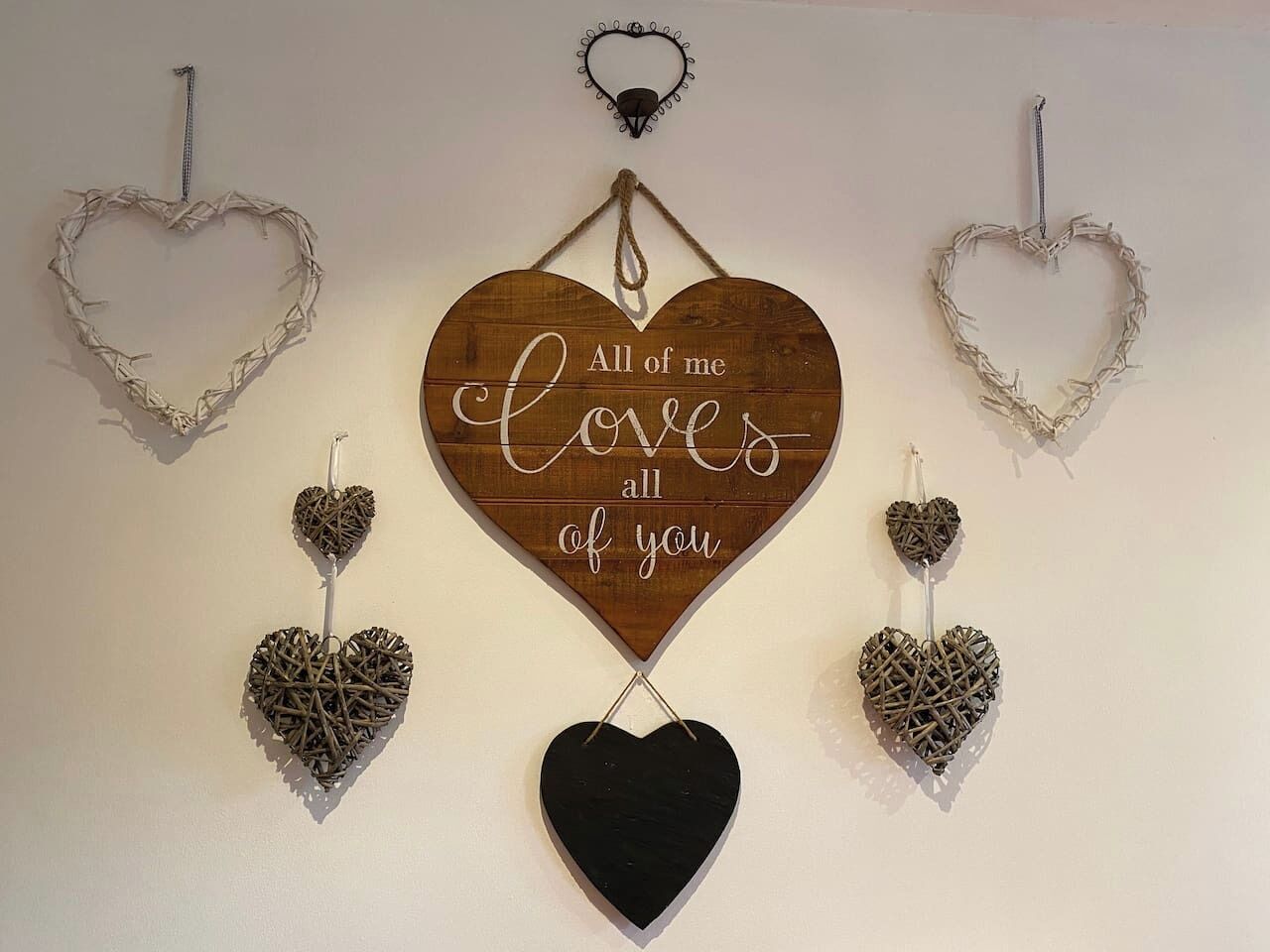 Un mur décoré de motifs en forme de cœur, comprenant une grande enseigne en bois au centre avec l’inscription « Je vous aime tous » et de plus petits cœurs réalisés dans différents matériaux.