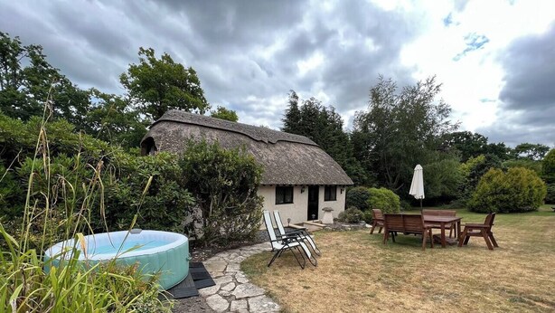 A rustic house with a thatched roof, a stone pathway leading to a wooden table and chairs, and a small swimming pool.
