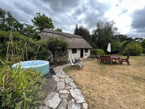 Una casa rústica con techo de paja, un sendero de piedra que lleva a una mesa y sillas de madera, y una piscina pequeña.