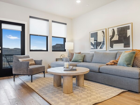 Un salón moderno con un sofá gris, una mesa de centro redonda y vistas al exterior a través de amplios ventanales.
