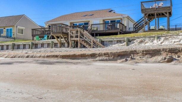 Een strandhuis met een houten terras, een trap naar een balkon en een zandstrand op de voorgrond.