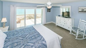 Een slaapkamer met een bed, een dressoir, een televisie, een schommelstoel en uitzicht op de oceaan door grote ramen.