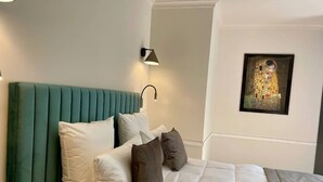 A bedroom with a teal upholstered headboard, white bedding, and a single wall-mounted light fixture. - Alfred Suites is an elegant, upscale residence ideally located in the vibrant he (Dijon)