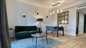A modern living room with a green sofa, a small round table, and a floor lamp. There is a dining area with a table and chairs, and a large window with a view. - Alfred Suites is an elegant, upscale residence ideally located in the vibrant he (Dijon)