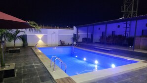 A swimming pool with a tiled deck, a palm tree, and a building with a white facade in the background.