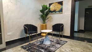 A lobby area with two black upholstered chairs, a gold-framed round table, a marble-patterned rug, a large potted plant, and a painting of a tree on the wall.
