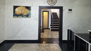 A hallway with a marble floor, a painting of a tree, a clock, and a staircase leading to a toilet.