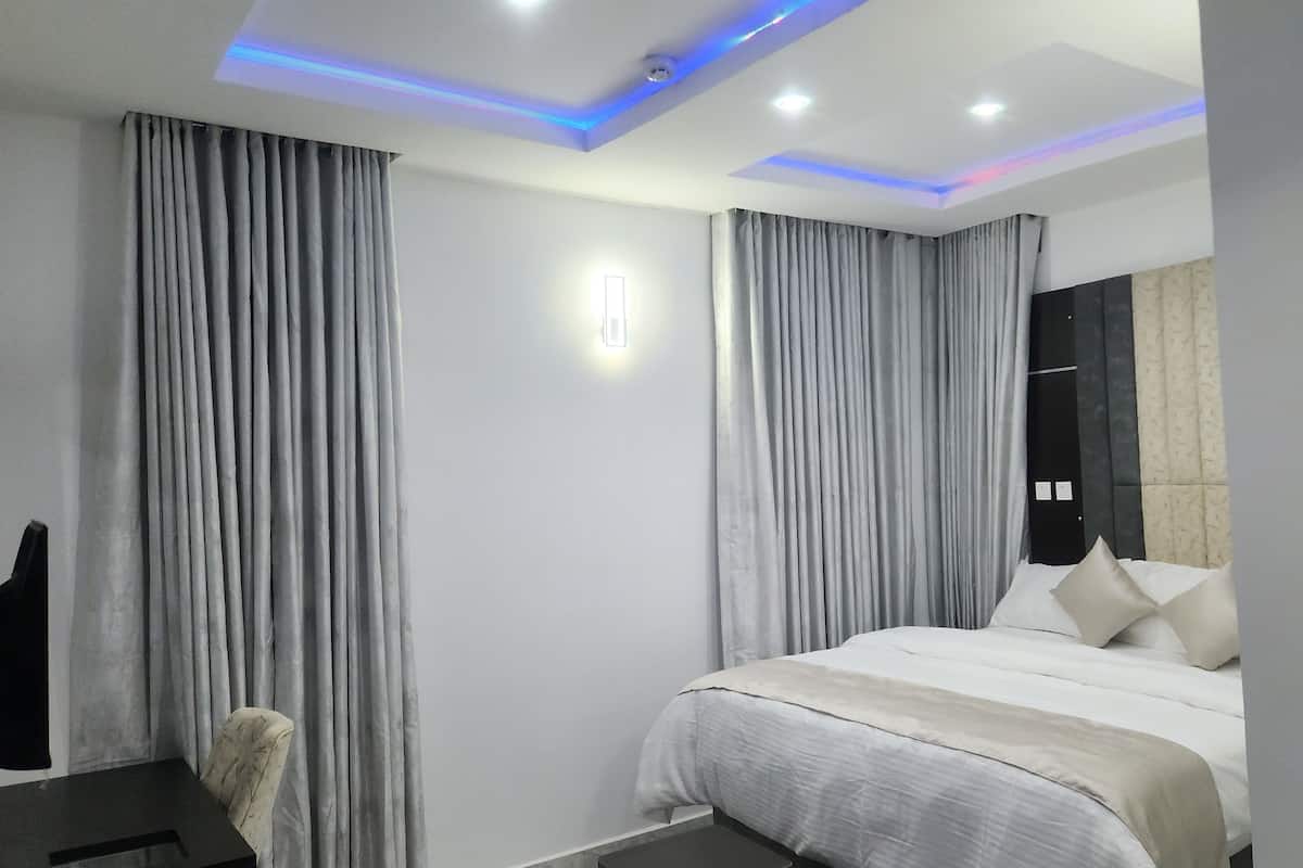 A modern hotel room with a large bed, grey curtains, a desk, and a chair.