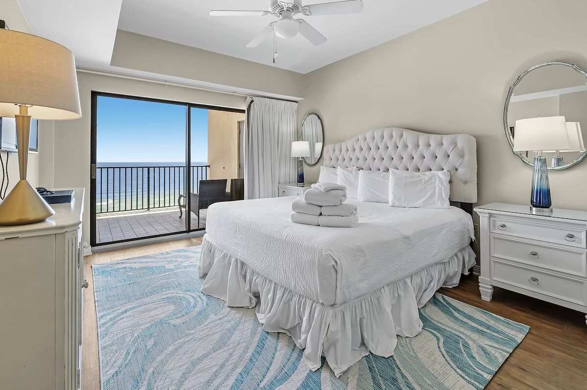 A hotel room with a large bed, a ceiling fan, a balcony with a view, and a white color scheme.