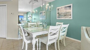 A dining area with a glass table, white chairs, teal walls, and a chandelier. - The Palms West 813 (Orange Beach)