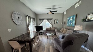 Ein modernes Wohnzimmer mit Essbereich, grauen Sofas, einem Couchtisch, einem Fernseher und einem Deckenventilator.