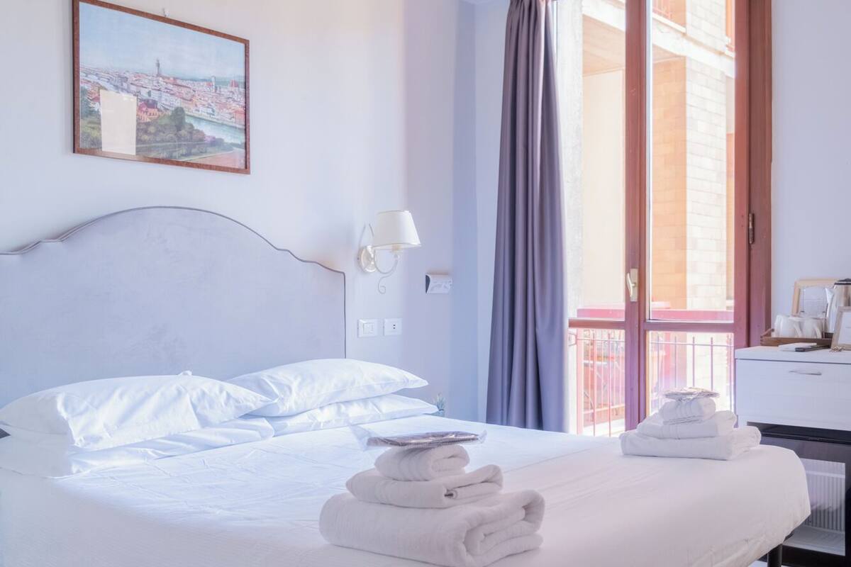 A neatly made bed with white linens and towels, a bedside lamp, a framed cityscape picture on the wall, and a wooden floor.