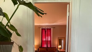 Een smalle gang die leidt naar een slaapkamer met een bed, nachtkastje, lamp en een raam met rode gordijnen.