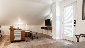 A cozy living room with a couch, a small table, a TV on a stand, and a door with blinds. - The Loft at Park Place Cozy Studio on the Park (Columbus)