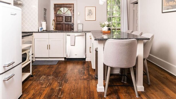 Una cocina con armarios blancos, piso de madera, zona de comedor con una mesa y sillas, y ventana con vista a follaje.