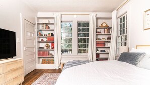 Un dormitorio con cama, televisión, estantería y ventana con cortinas.