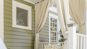 A covered porch with a wicker sofa and a bench, a window with curtains, and a white railing. - Historic Park Place Retreat  3 Bdrm on the Park (Columbus)