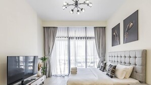 A modern bedroom with a large bed, a TV on a wooden stand, and a view of the outdoors through sheer curtains. - Relaxing Studio Retreat in Vibrant Meydan City, (Dubai)