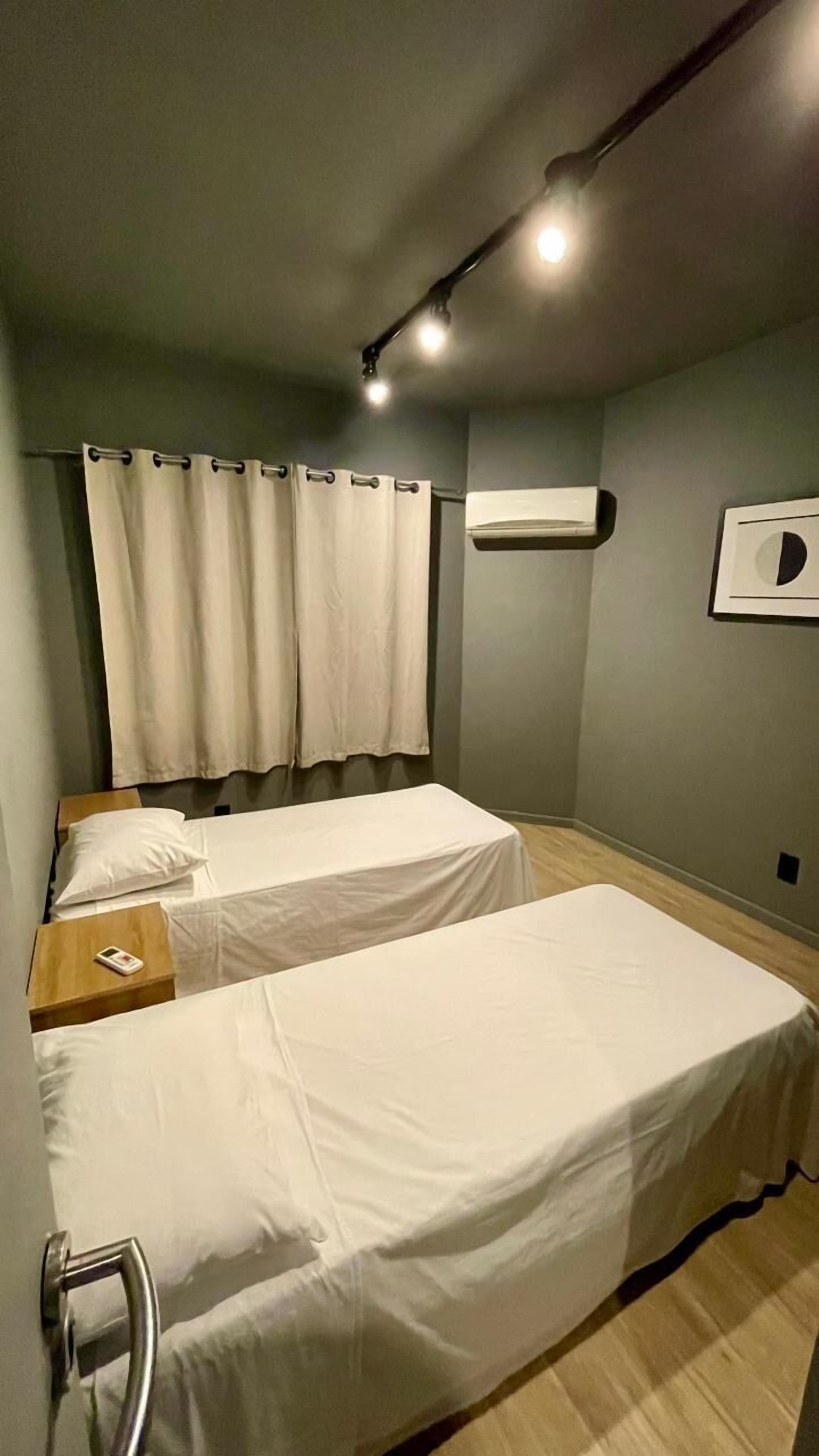 A small, dimly lit room with two single beds, a wooden nightstand, a wall-mounted air conditioner, and a curtain.
