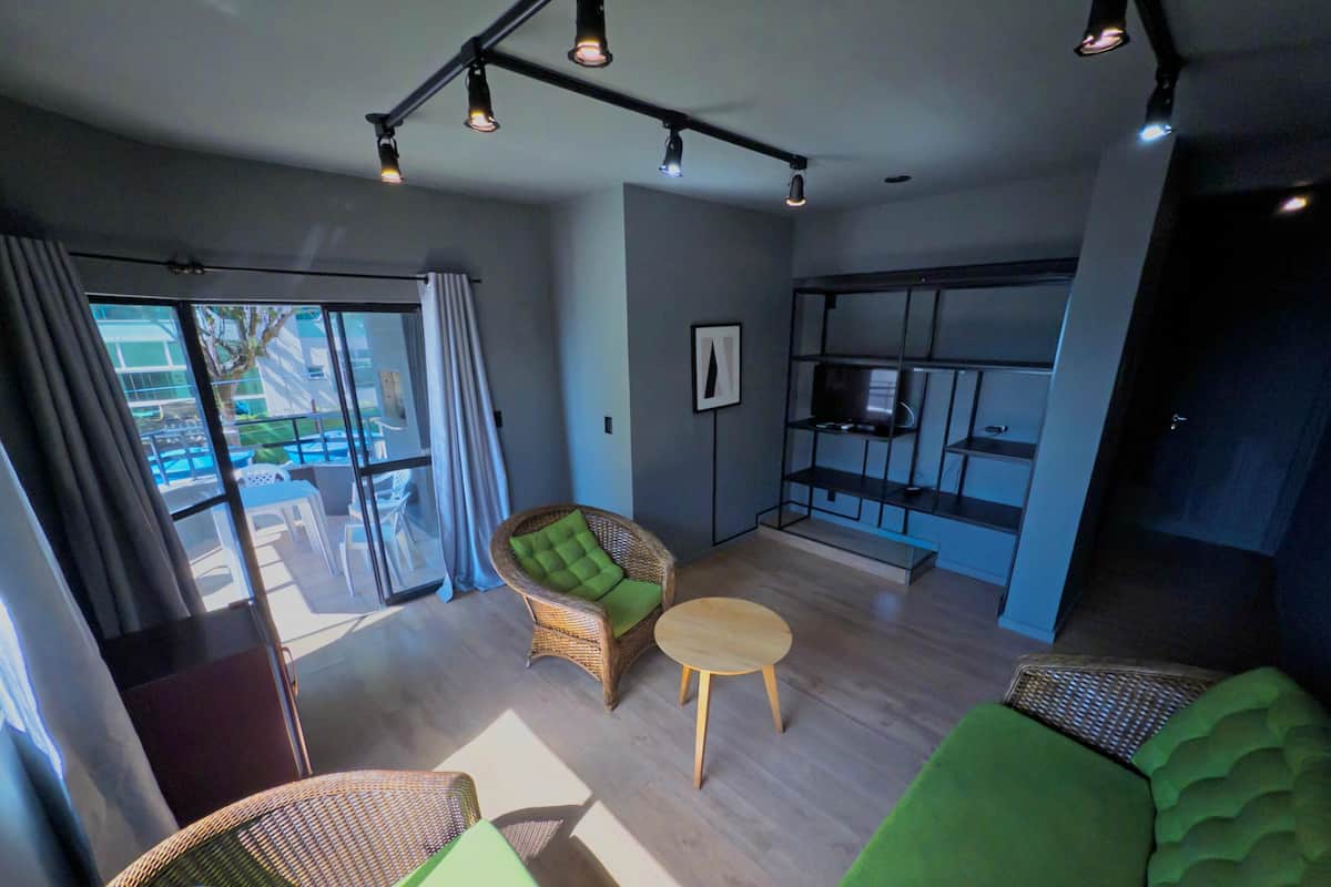A modern living room with a green sofa, wicker chairs, a small round table, and a kitchen area with a TV and shelves.