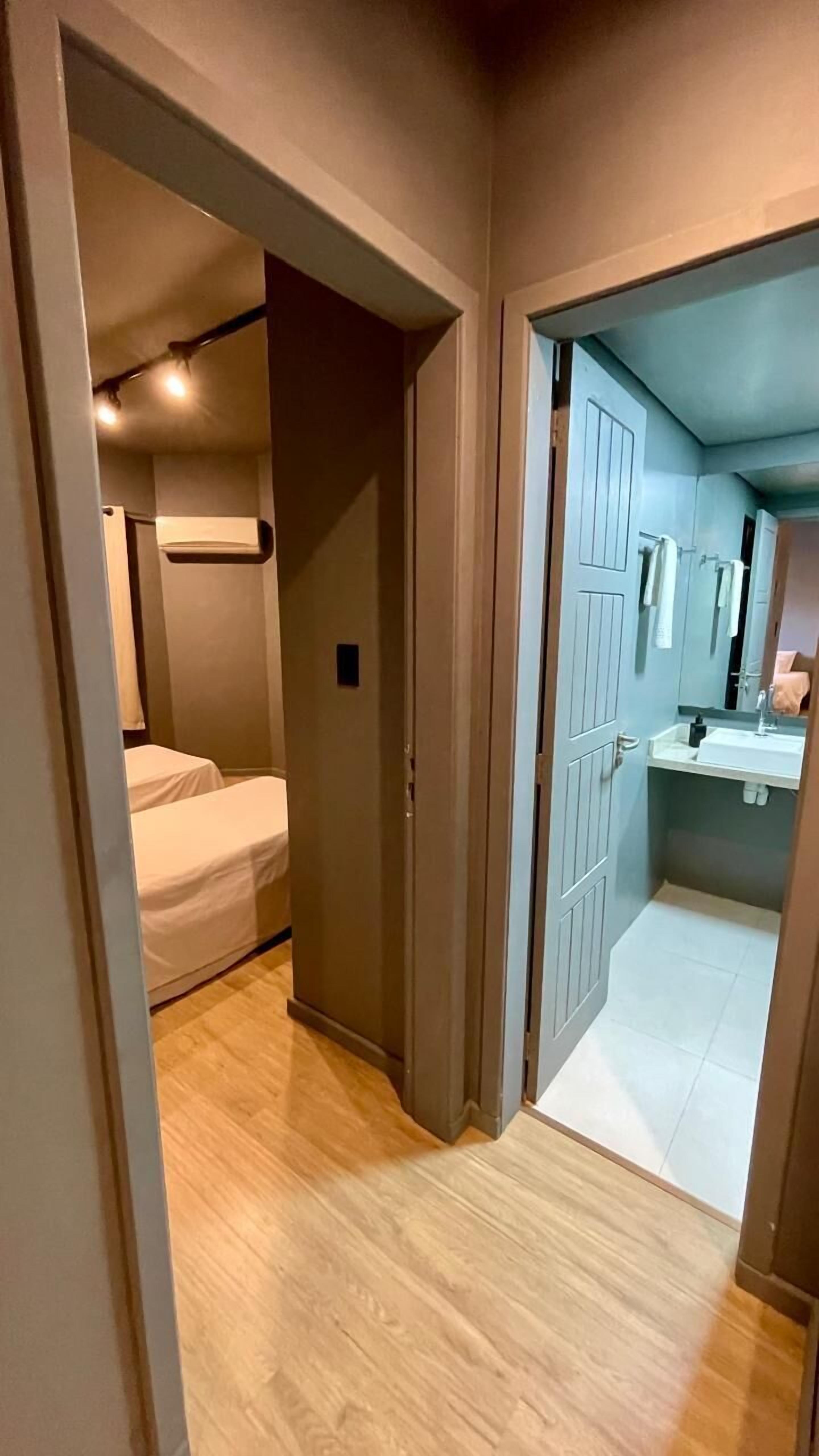 Un pasillo que conduce a un dormitorio y un baño. El dormitorio tiene una cama y un estante pequeño. El baño tiene un lavabo y un espejo.