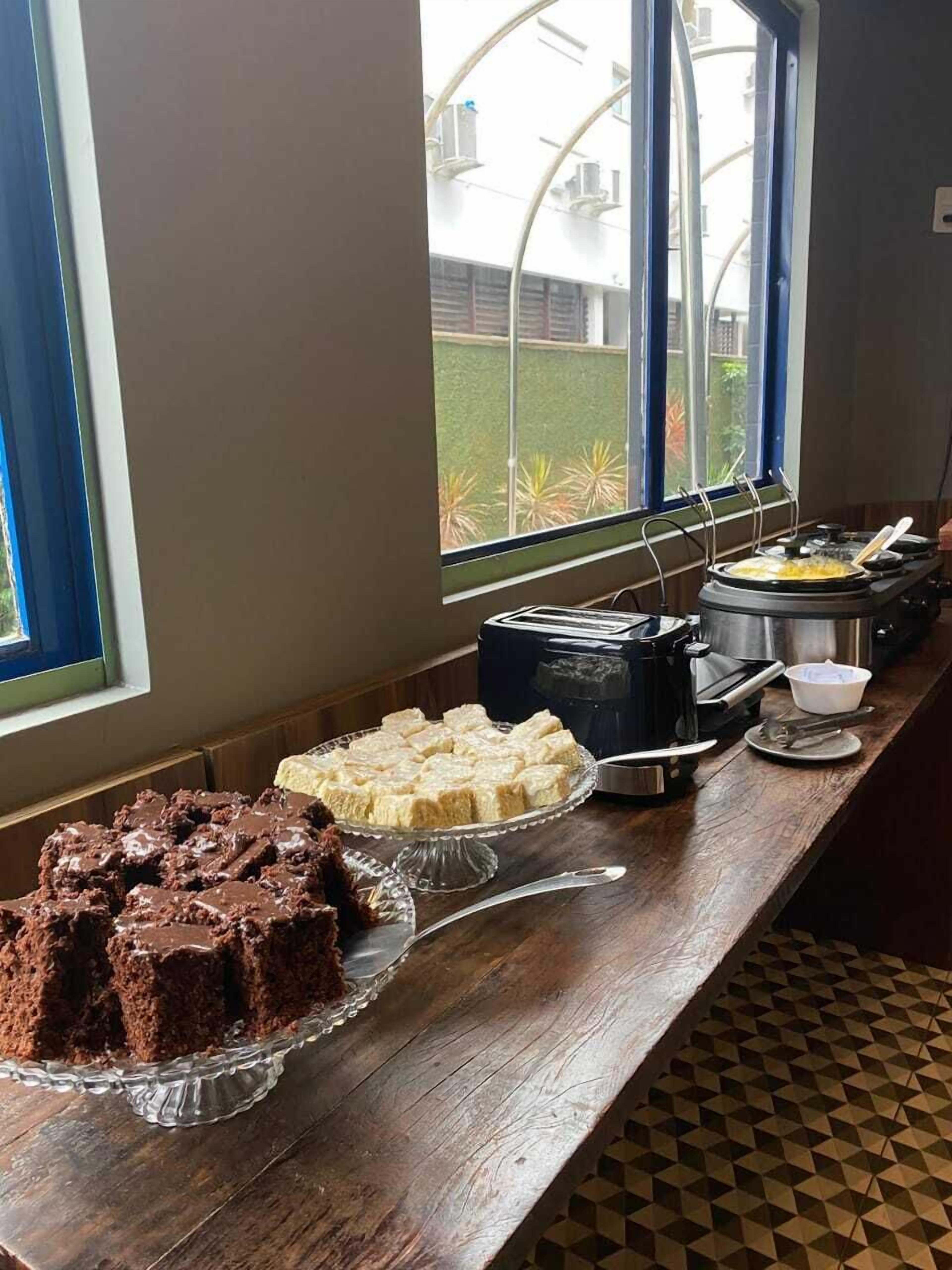 A buffet table with various desserts, including brownies and a cake, in a room with large windows and a view of a building outside.