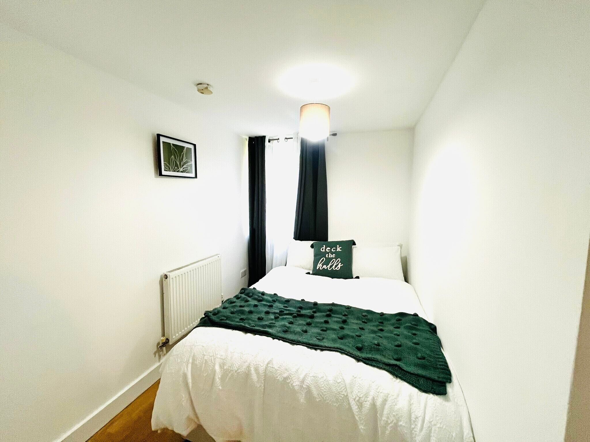 A bedroom with a bed, a radiator, a framed picture on the wall, and a pillow with text.