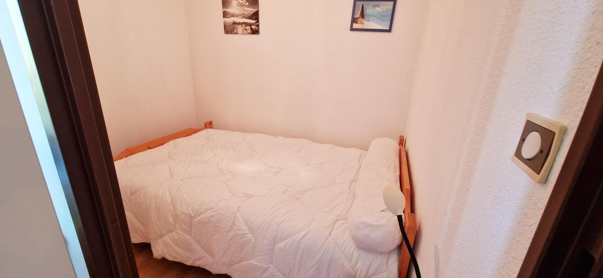 A small, neatly made bed in a compact room with a plain wall and a door visible.