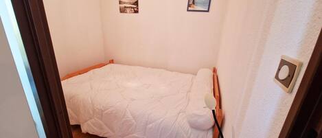 A small, neatly made bed in a compact room with a plain wall and a door visible.