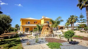 A yellow villa with a large rock centerpiece, surrounded by a gravel courtyard and palm trees. - Quinta do Sol in Guia (Albufeira)