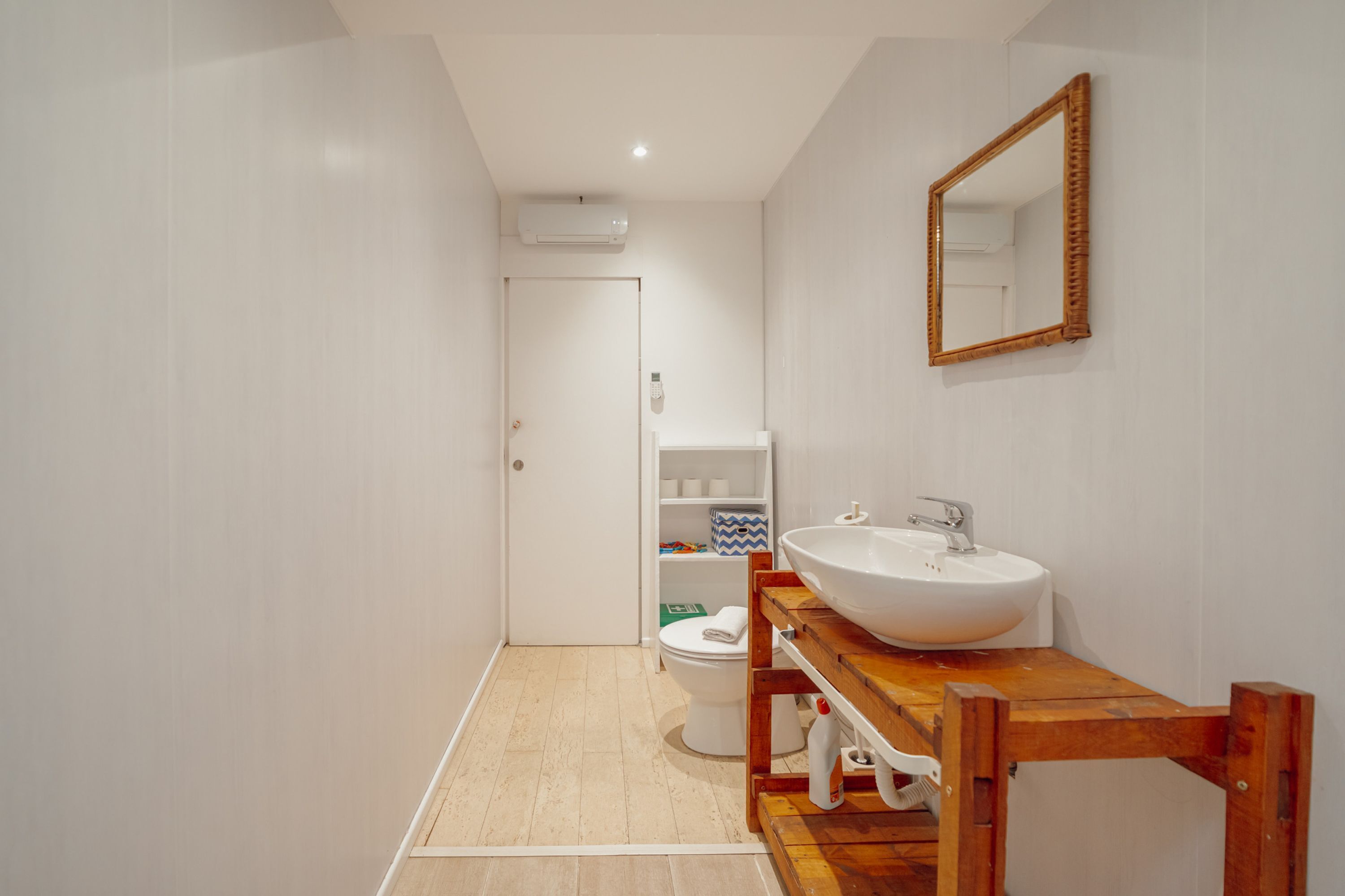 A bathroom with a wooden vanity, white sink, and a mirror. There is a toilet and a shelf with toiletries.