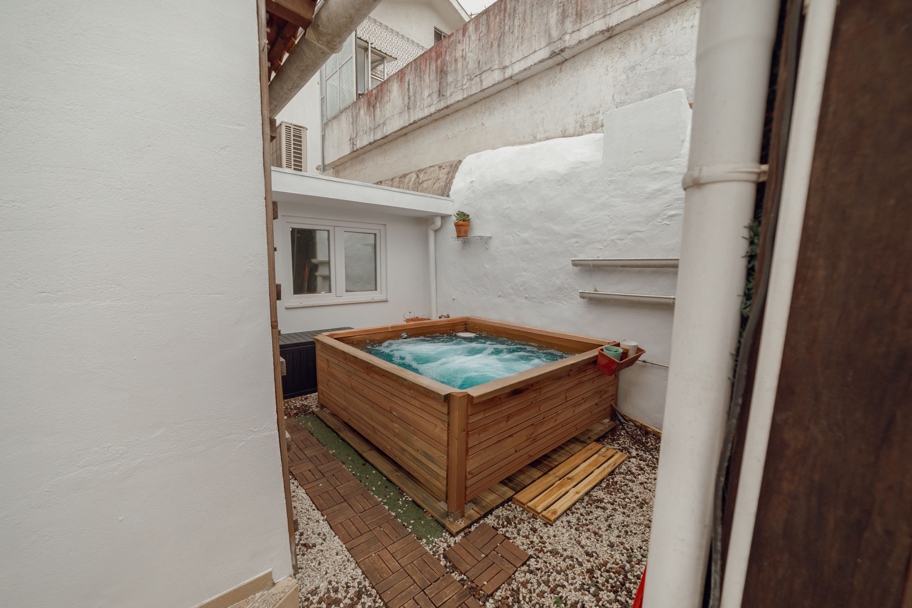 A wooden hot tub in a narrow outdoor space with a white wall and a window.