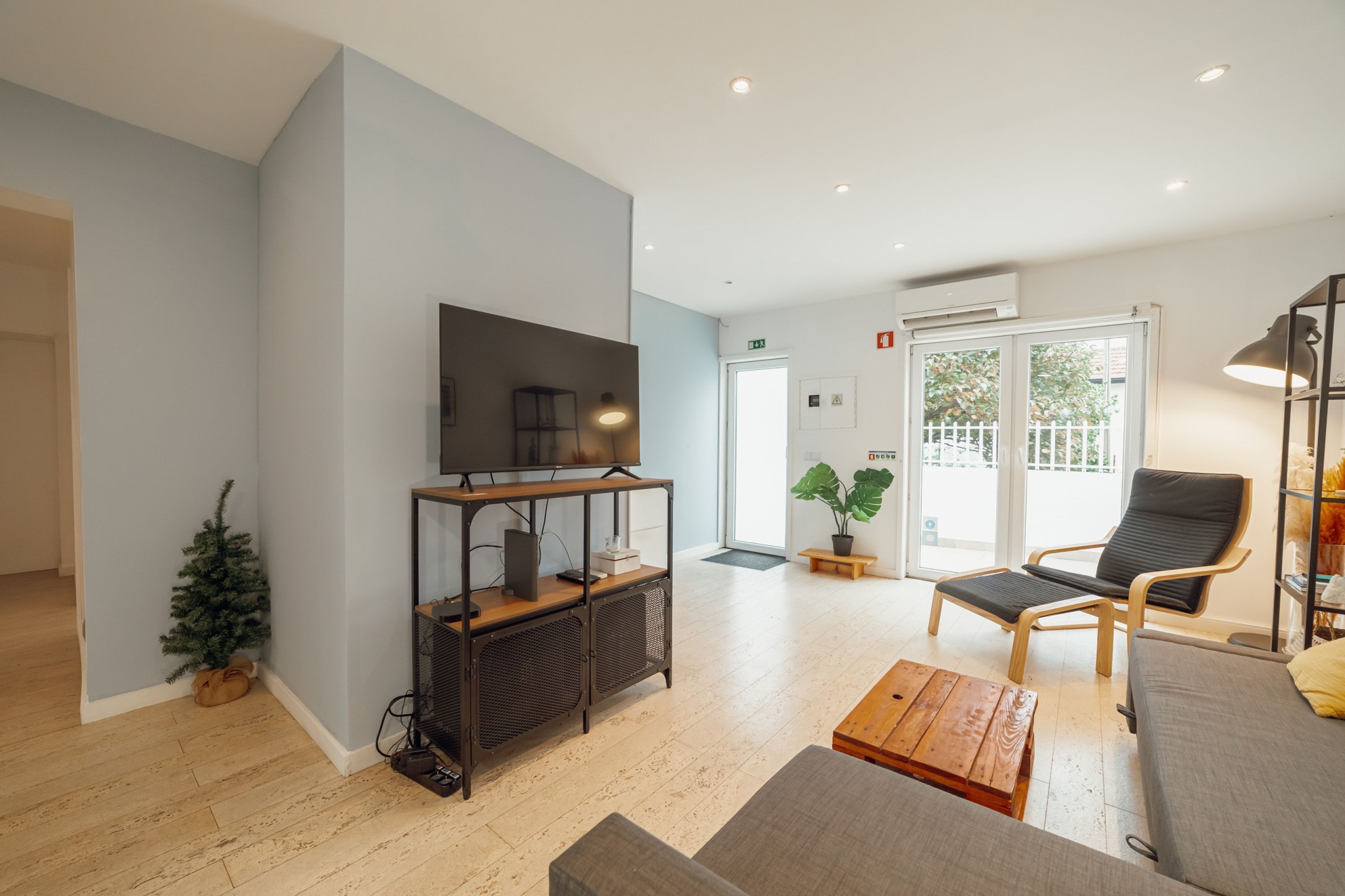 Una sala de estar moderna con un televisor de pantalla plana sobre un soporte de madera, un pequeño árbol de Navidad, un sofá, una mesa de centro y una silla con una planta al lado de la puerta.