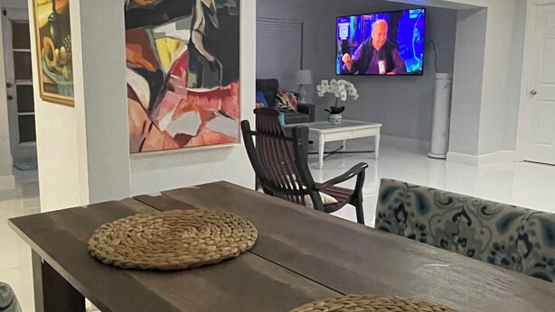 A modern living room with a wooden dining table, woven placemats, patterned cushions, a television, and abstract art on the walls. - Enjoy SWFL (Fort Myers)