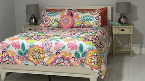 A bedroom with a floral bedspread, two bedside lamps, and a painting of red flowers on the wall. - Enjoy SWFL (Fort Myers)