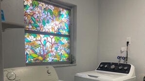 A laundry room with a front-loading washer and dryer, a window with a stained glass design, and a wall-mounted electrical outlet. - Enjoy SWFL (Fort Myers)