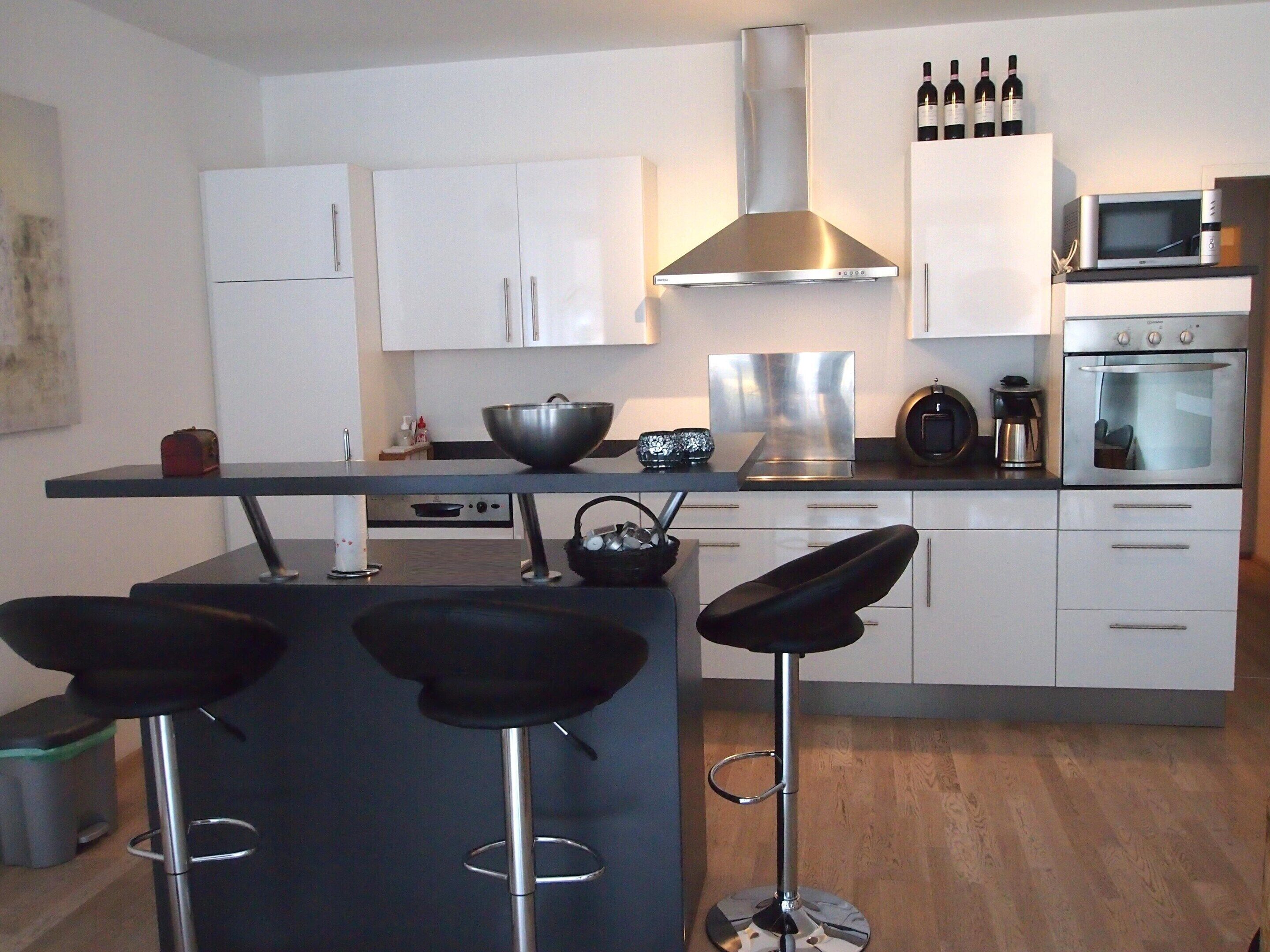 A modern kitchen with white cabinets, a stainless steel range hood, a microwave, an oven, and a bar area with stools.