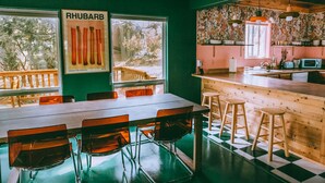 Een restaurant met een houten tafel en stoelen, een keukenhoek met een aanrecht en barkrukken, en een poster van rabarber aan de muur.